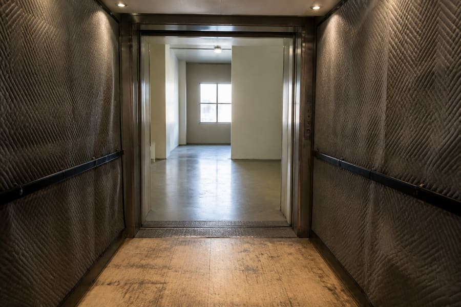 A building freight elevator hung with quilted protective pads, doors open onto a plain corridor